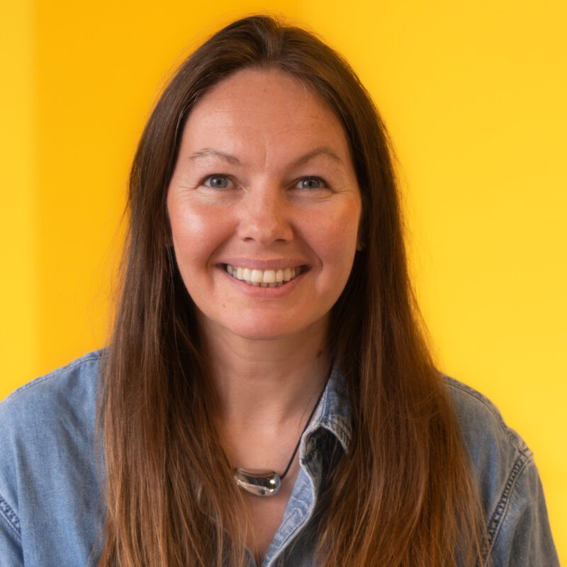 A woman with long brown hair smiles at the camera. She is wearing a denim shirt and a silver necklace, standing in front of a bright yellow background.