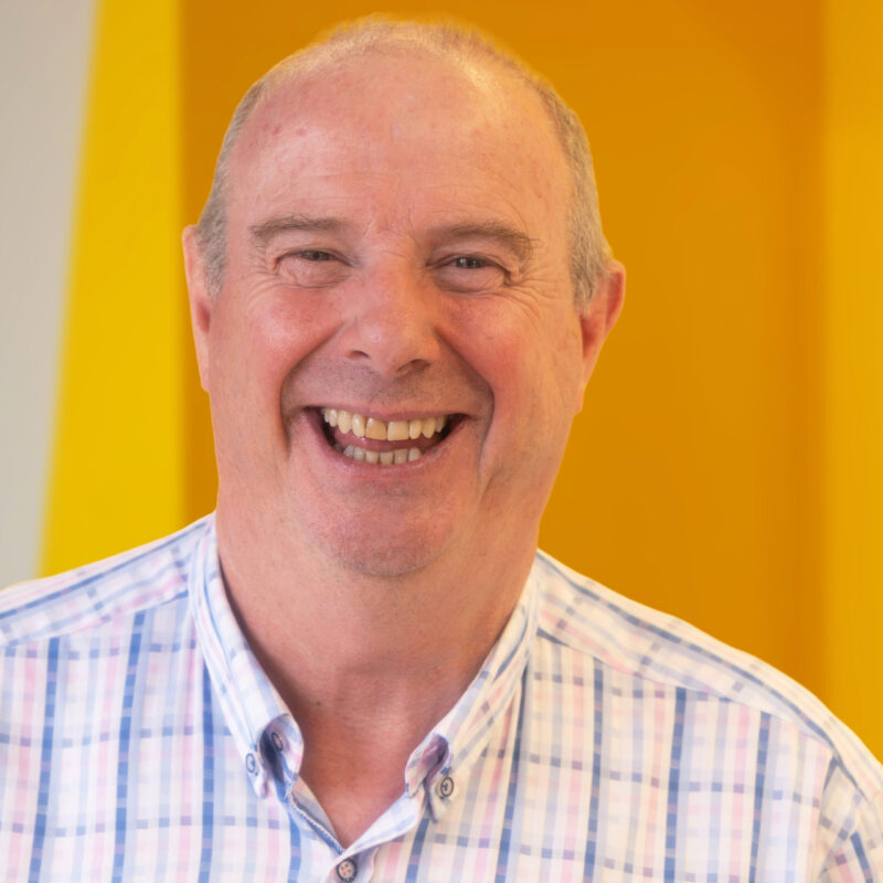 A middle-aged man with short hair and a checked shirt smiles broadly in front of a bright yellow background.