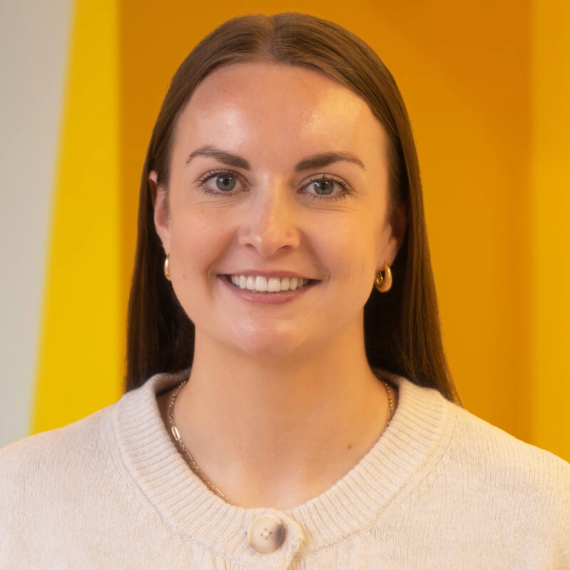 A woman with straight brown hair, wearing a cream jumper, gold hoop earrings, and a gold necklace, smiles in front of a bright yellow background.