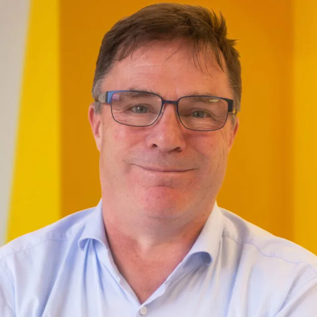 A middle-aged man with short brown hair, glasses, and a light blue shirt smiles softly in front of a bright yellow background.
