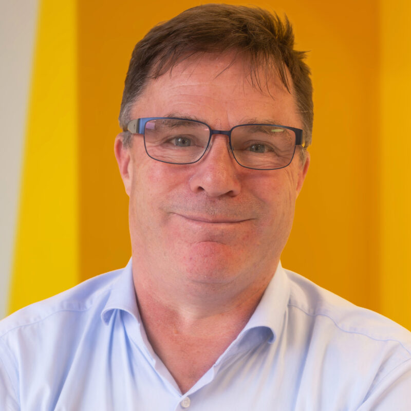 A middle-aged man with short brown hair, glasses, and a light blue shirt smiles softly in front of a bright yellow background.
