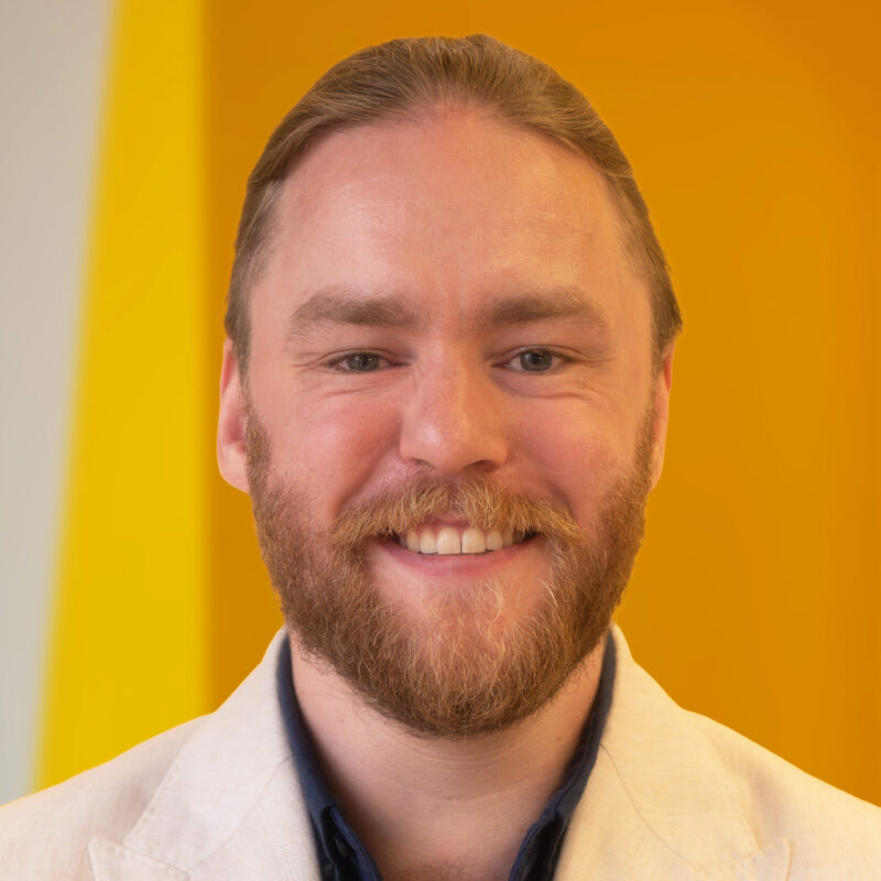 A smiling man with a beard wearing a light-coloured jacket and dark shirt stands in front of a bright yellow and orange background.