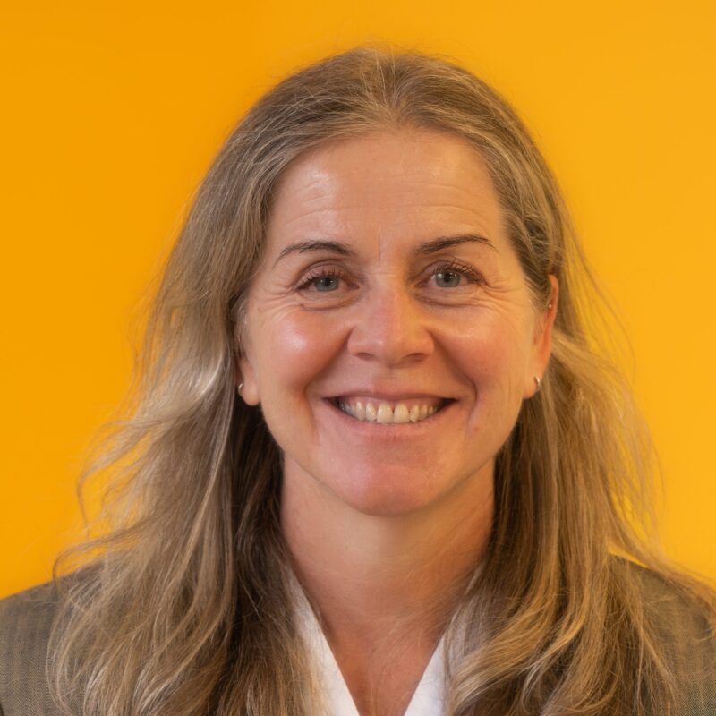 A woman with long, light brown hair smiles at the camera in front of a bright yellow background. She wears a white top and a grey blazer.
