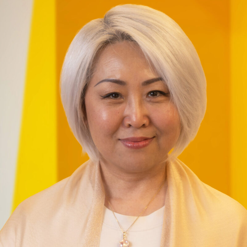 A woman with short, light blonde hair smiles gently, standing in front of a bright yellow background. She wears a cream-coloured top and a necklace.